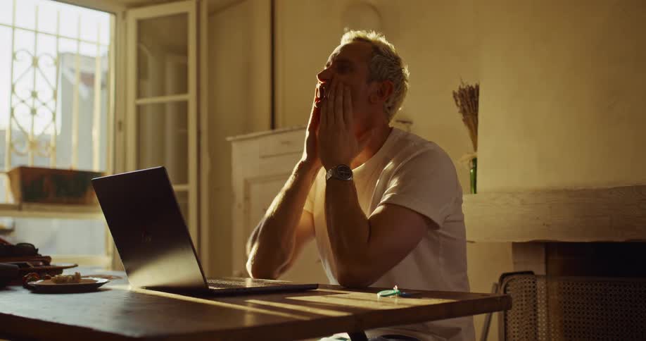 A stressed man with bleached blond hair has an emotional and expressive conversation on a video call from his rustic, sunlit home