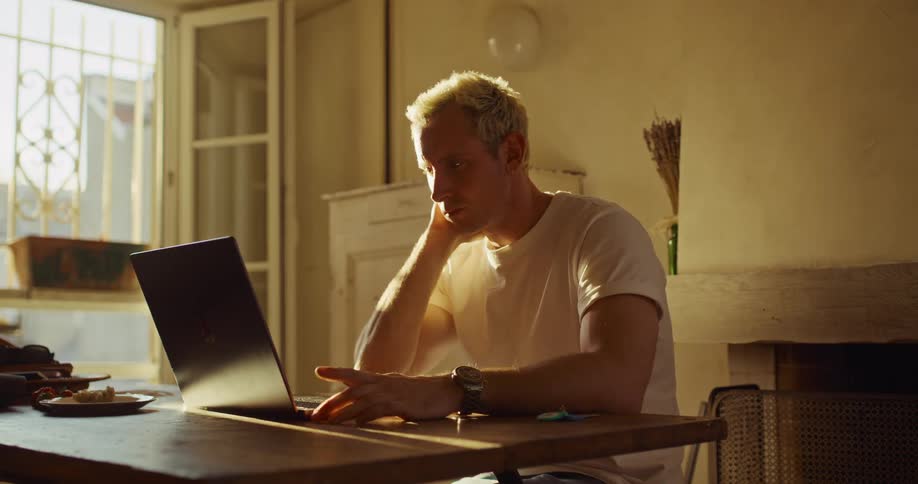 A thoughtful man working on his laptop in a sunlit room rubs his tired face before continuing to type