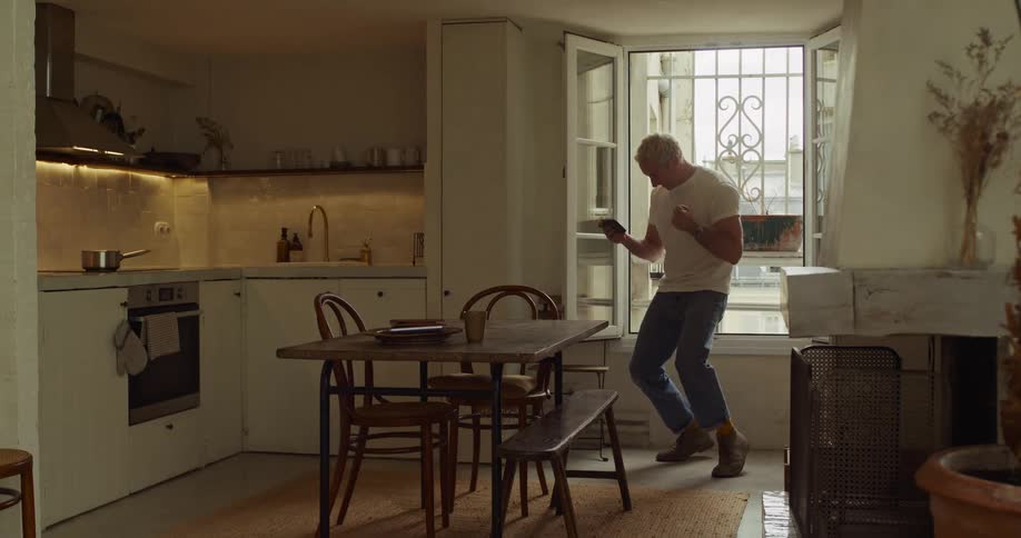 A happy man dances in his rustic apartment kitchen after getting great news on his smartphone, then starts to cook