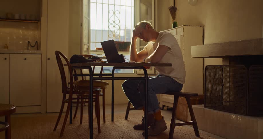 A thoughtful young man with bleached blonde hair works from home on his laptop in a sunlit room, pausing to stretch