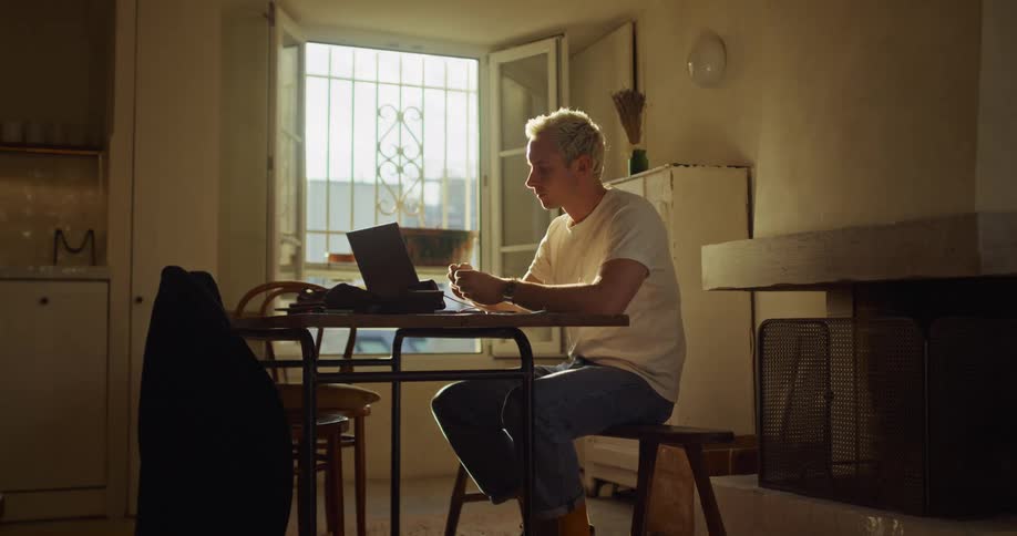 Young man with bleached hair works from home on his laptop, sitting at a table in a rustic, sun-drenched room