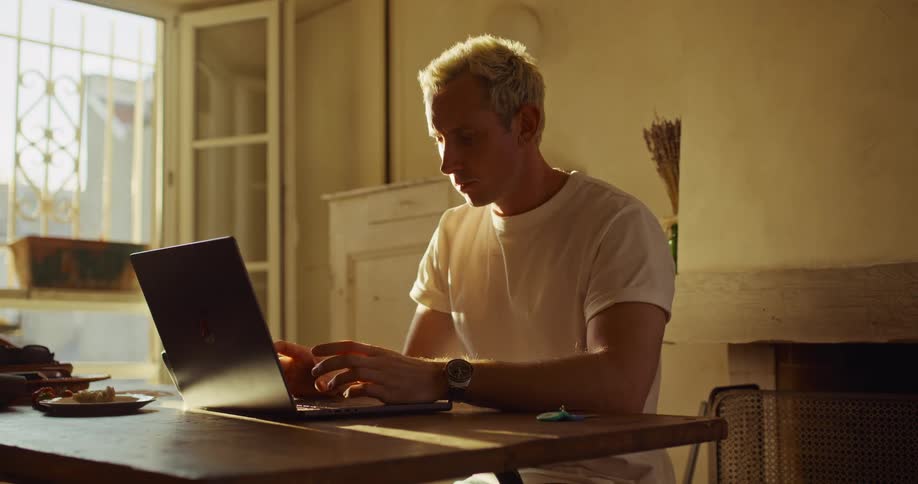 A pensive man with blond hair works on his laptop in a warmly lit room, showing frustration before continuing to type