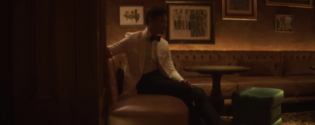 An elegantly dressed man in a white tuxedo sits alone and looks off-camera in a dimly lit, upscale lounge