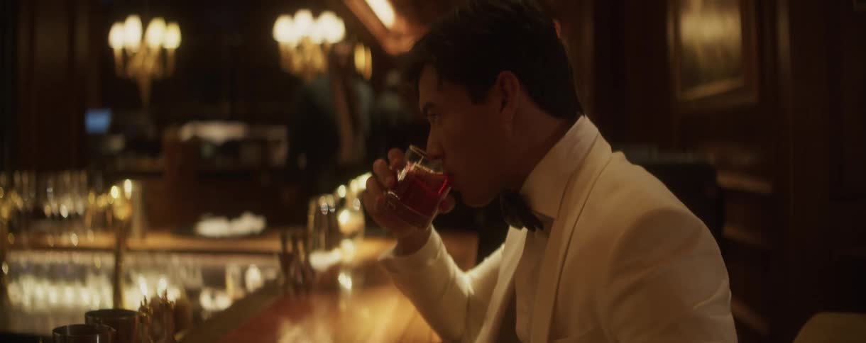 A sophisticated man in a white tuxedo takes a slow sip of a red cocktail at an elegant bar