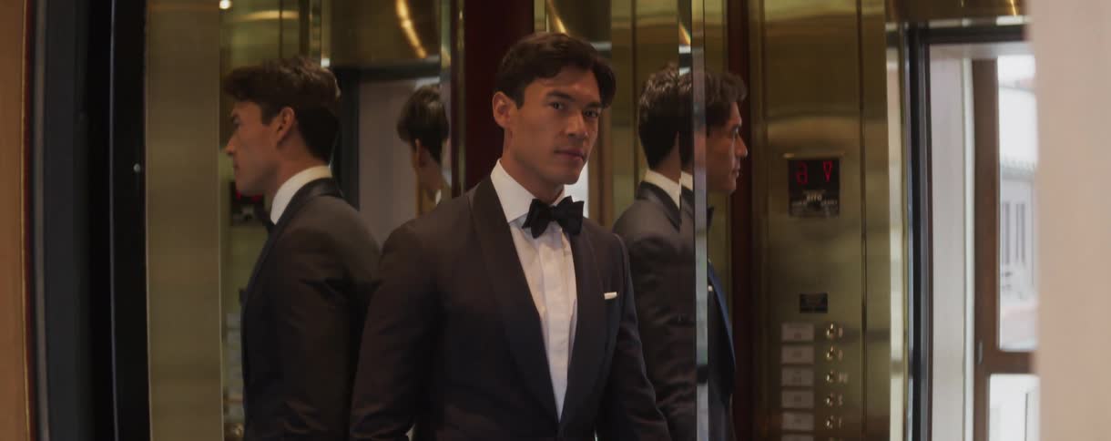 An elegant man in a tuxedo smiles slightly as the doors of a luxurious, mirrored elevator open