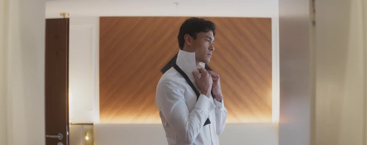 A well-dressed young man carefully adjusts his crisp white shirt collar in a stylish room