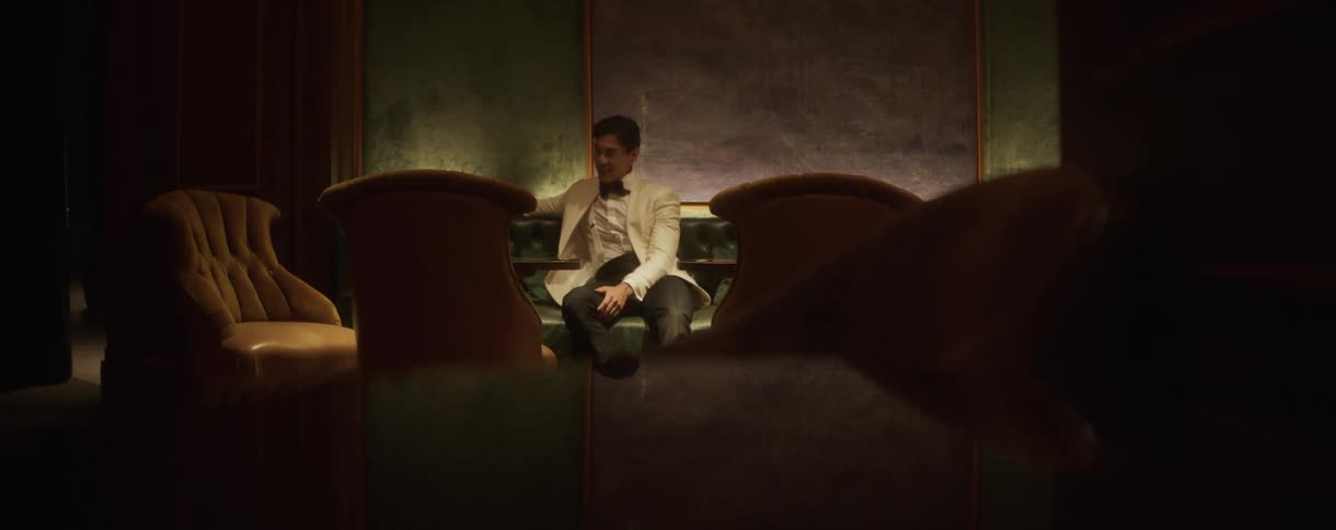 An elegant man in a white dinner jacket sits expectantly in a dimly lit, luxurious lounge booth