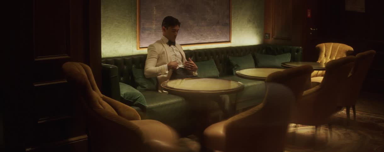 A dapper man in a white tuxedo waits alone in a sophisticated, dimly lit lounge