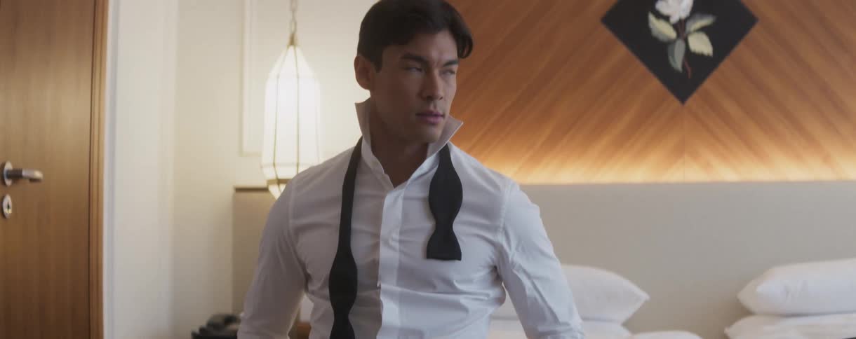 A handsome man in a tuxedo shirt and untied bow tie sits on a hotel bed and looks intently at the camera
