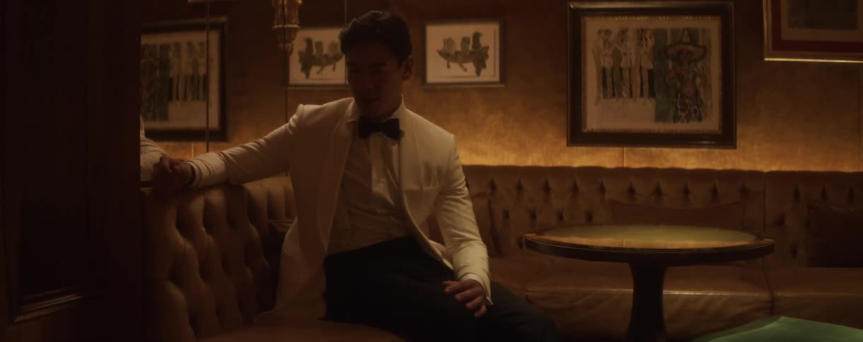 A handsome man in a formal white tuxedo waits alone in a sophisticated, moody lounge
