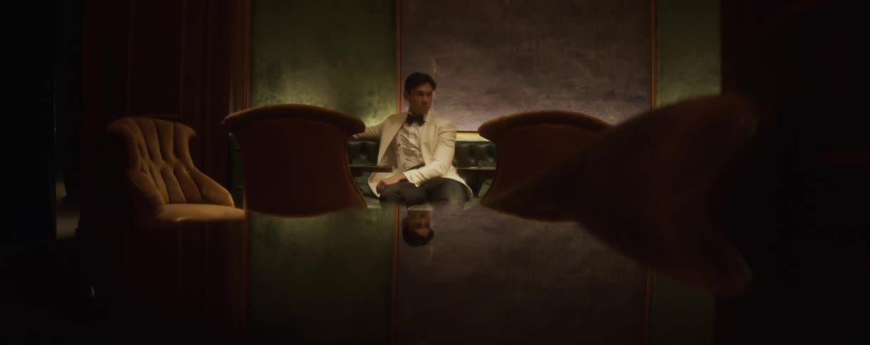 A stylish man in a white tuxedo waits alone at a reflective table in a dimly lit, luxurious lounge