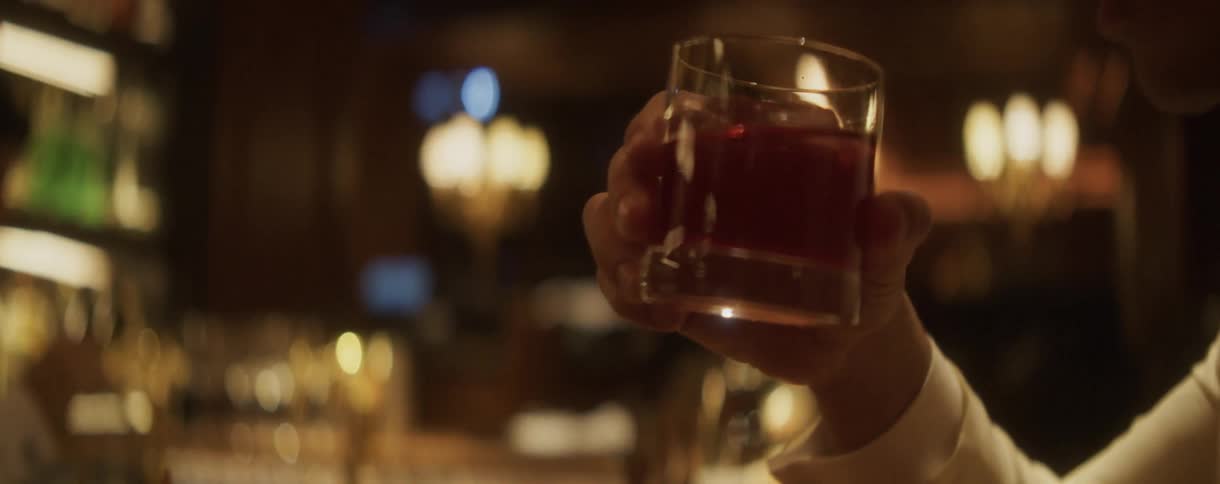 A well-dressed man slowly sips a red cocktail from a glass at an elegant, dimly-lit bar