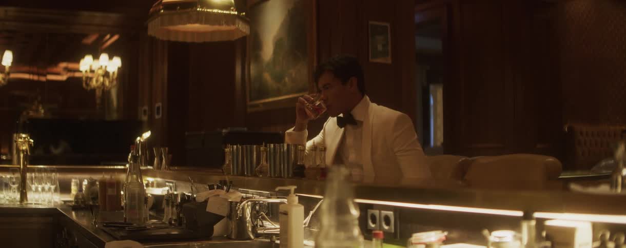 A dapper man in a white tuxedo sips a cocktail alone at a dimly lit, elegant bar