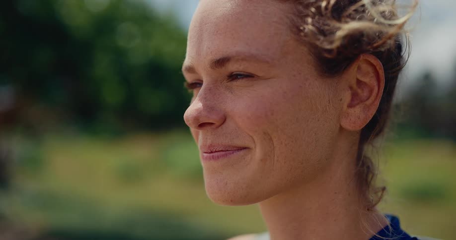 A serene woman with freckles and curly hair smiles peacefully outdoors as the wind gently blows