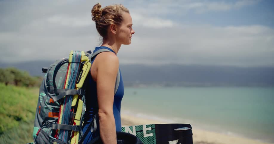 A smiling female kitesurfer stands with her board and gear on a sandy dune, looking out at the ocean before her session