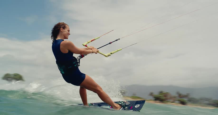 An athletic woman skillfully kitesurfs away from a tropical shore, captured in slow motion from an immersive water-level view
