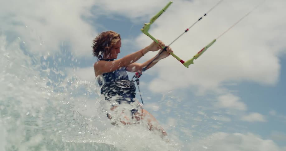 A woman kitesurfs across the ocean in slow motion, splashing water towards the camera on a sunny day