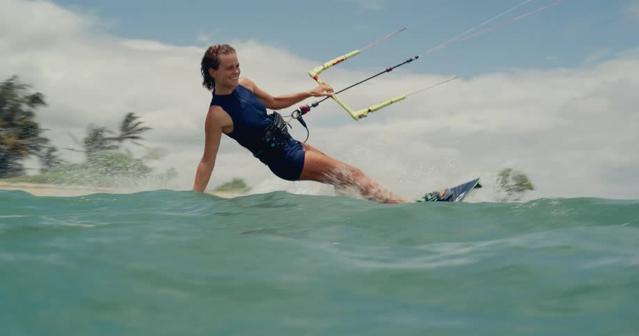 A happy young woman kitesurfs through the ocean waves in slow motion, shot from the water with a tropical beach in the background