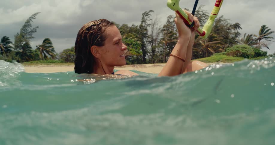A happy young woman kitesurfer smiles while floating in the ocean holding her kite bar in slow motion