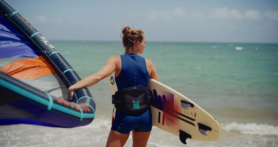 A woman kiteboarder with her gear stands at the edge of the water looking out at the ocean on a sunny day