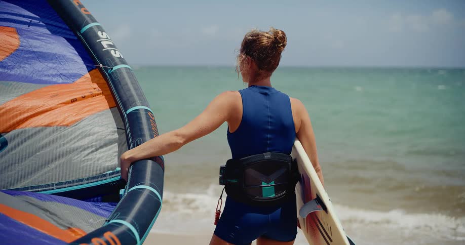 A female wing foiler in a blue wetsuit stands on a sunny beach, holding her board and wing while looking out at the ocean