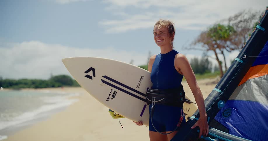 A confident and happy female kitesurfer stands on a sunny tropical beach with her board and kite, ready for an adventure