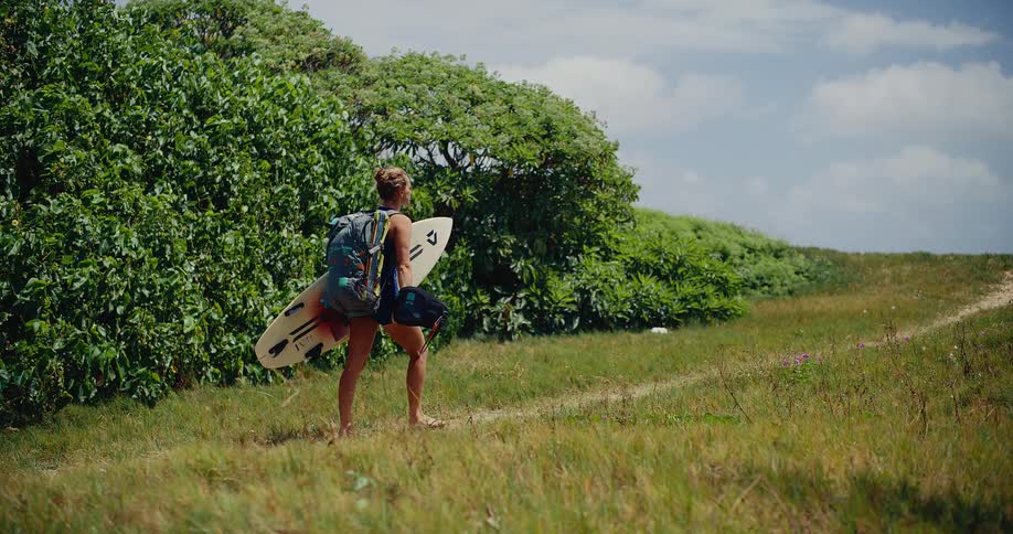 Slow motion shot of a female surfer carrying a surfboard and backpack, walking along a grassy coastal path on a sunny day