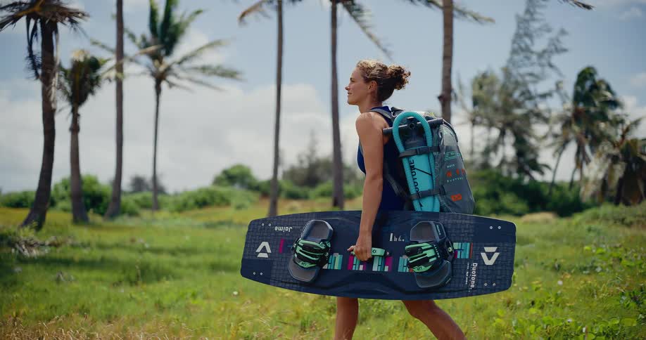 A happy young woman carries her kiteboard and gear while walking through a tropical landscape of grass and palm trees