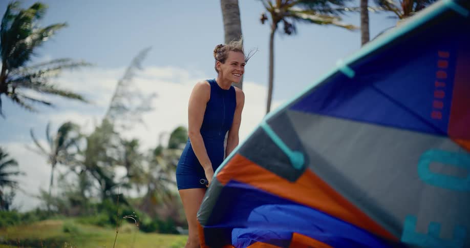 A smiling female kitesurfer in a wetsuit pumps up her colorful kite on a windy, tropical beach with palm trees