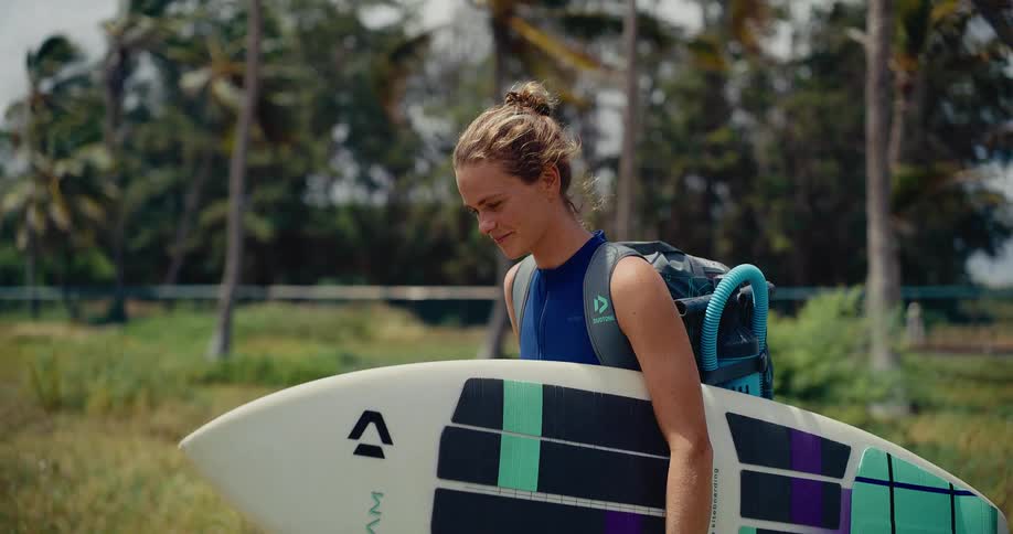 A smiling young woman in a wetsuit carries her kiteboard and backpack while walking through a windy, tropical paradise