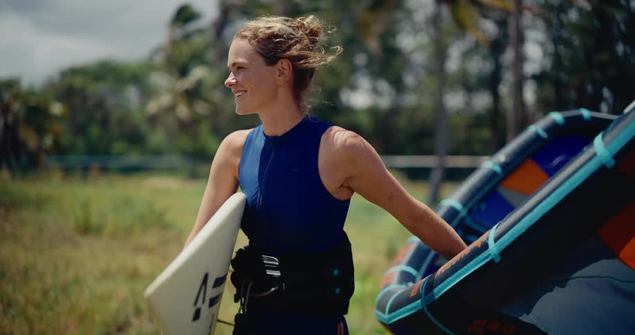 A smiling female athlete confidently carries her kitesurfing board and kite while walking through a tropical, windy landscape
