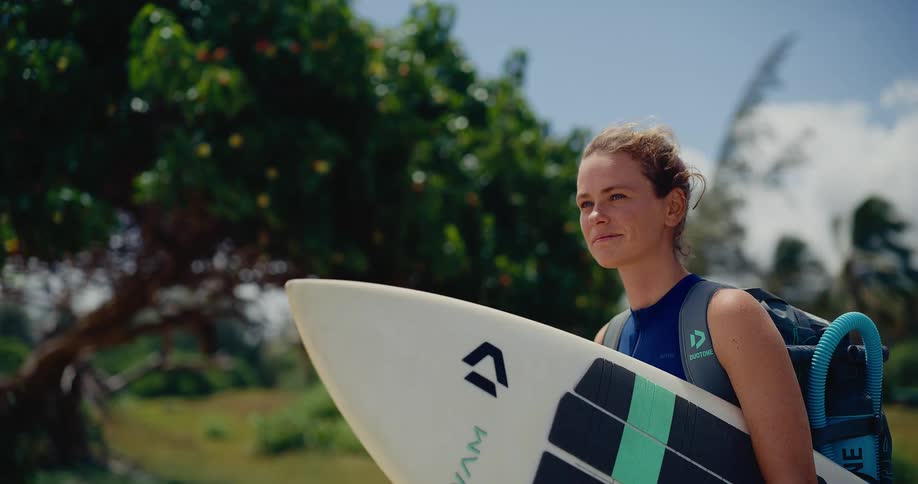 A confident female kitesurfer in a wetsuit walks with her surfboard on a windy tropical day in slow motion