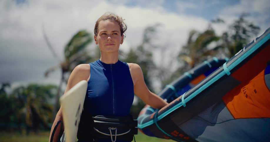 A confident female kitesurfer stands with her board and kite on a windy tropical beach, ready for an adventure