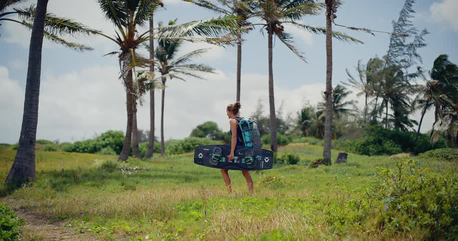 A young woman carries her kitesurfing board and backpack while walking through a windy, tropical field of palm trees