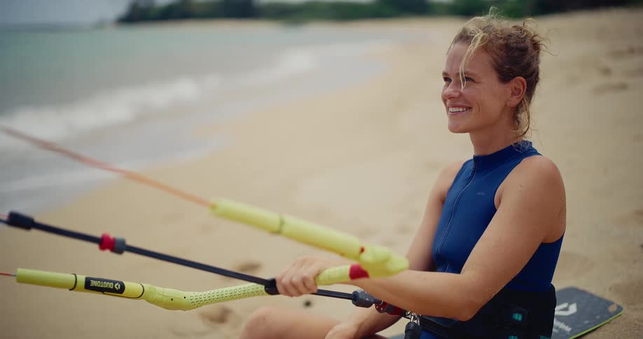A happy young woman sits on a sandy beach with her kitesurfing gear, smiling as she looks out at the ocean