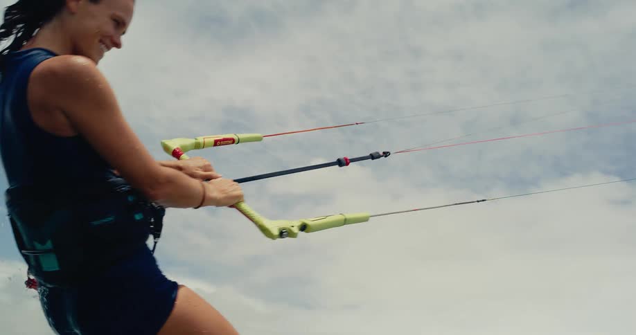 A smiling woman begins to kitesurf in the ocean, rising from the water and riding away from a tropical beach in slow motion