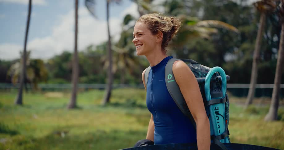 A smiling woman in a wetsuit walks with her kitesurfing equipment through a grassy field with palm trees on a windy day in slow motion