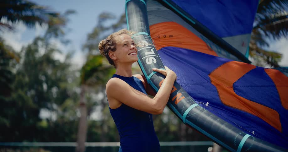 A smiling young woman in a swimsuit prepares her kitesurfing gear on a sunny, windy day with palm trees in the background
