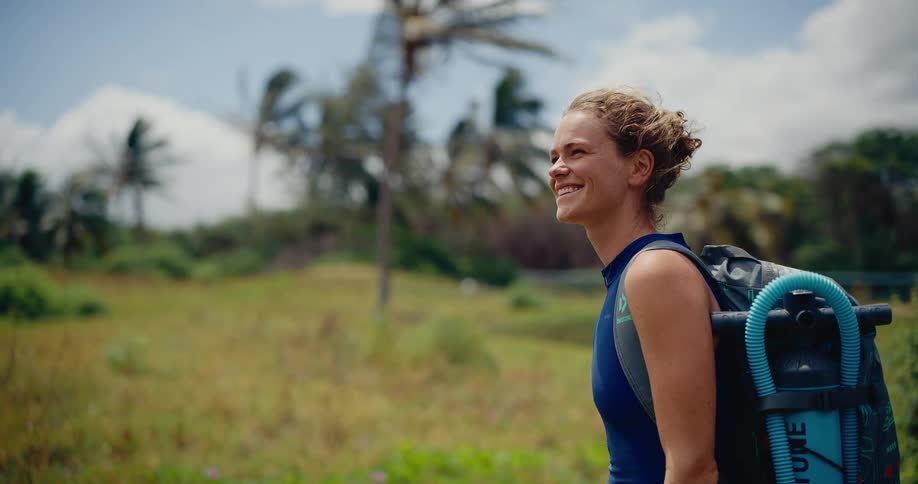 A smiling female adventurer with a backpack and water sports equipment enjoys the windy day in a lush tropical setting