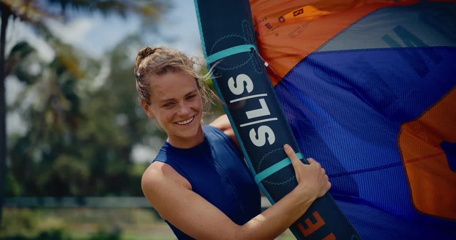A smiling female kitesurfer in a blue wetsuit happily prepares her kite equipment on a sunny tropical beach