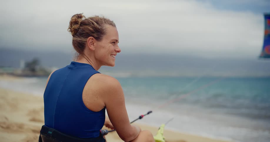Portrait of a smiling female kitesurfer sitting on the beach looking out at the ocean
