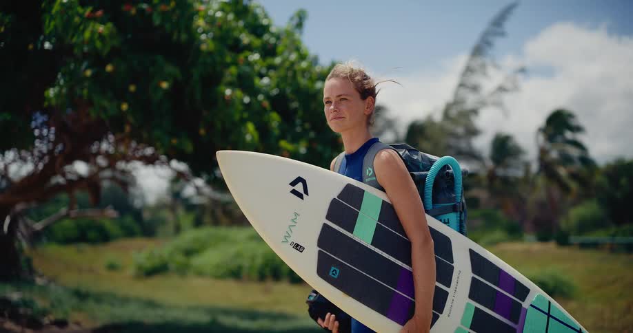Cinematic slow motion of a female kitesurfer in a blue wetsuit holding her board on a windy tropical coast