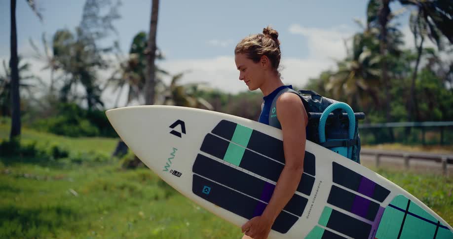 An athletic woman smiles as she carries her surfboard and kitesurfing gear while walking through a lush tropical landscape in slow motion