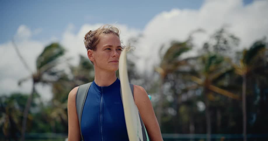A happy female kitesurfer in a wetsuit holds her board on a windy tropical beach, smiling as she looks out towards the horizon