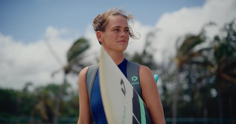 A smiling female surfer in a wetsuit holds her surfboard on a windy tropical beach as the wind blows through her hair in slow motion