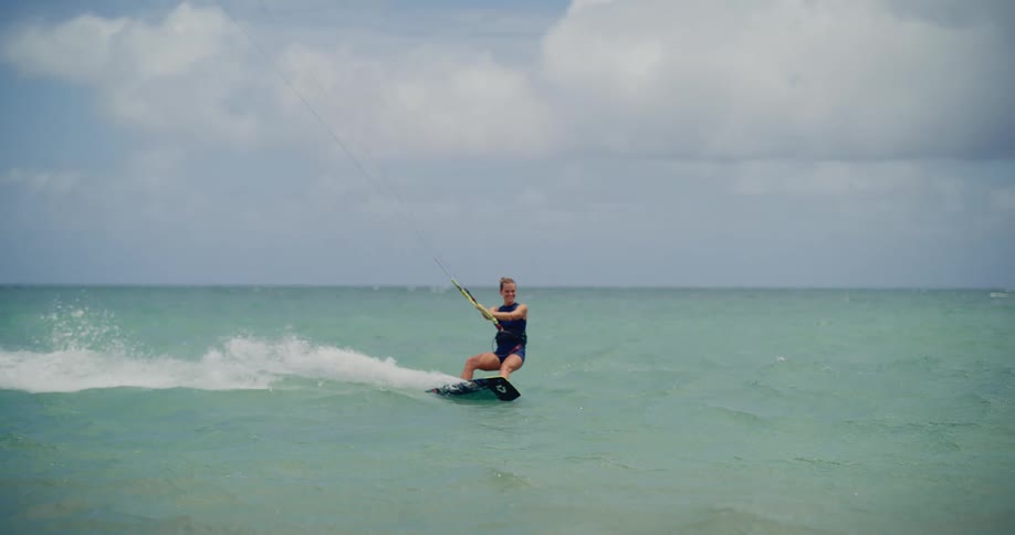 A female kitesurfer skillfully rides the turquoise ocean waves on a partly cloudy day in slow motion