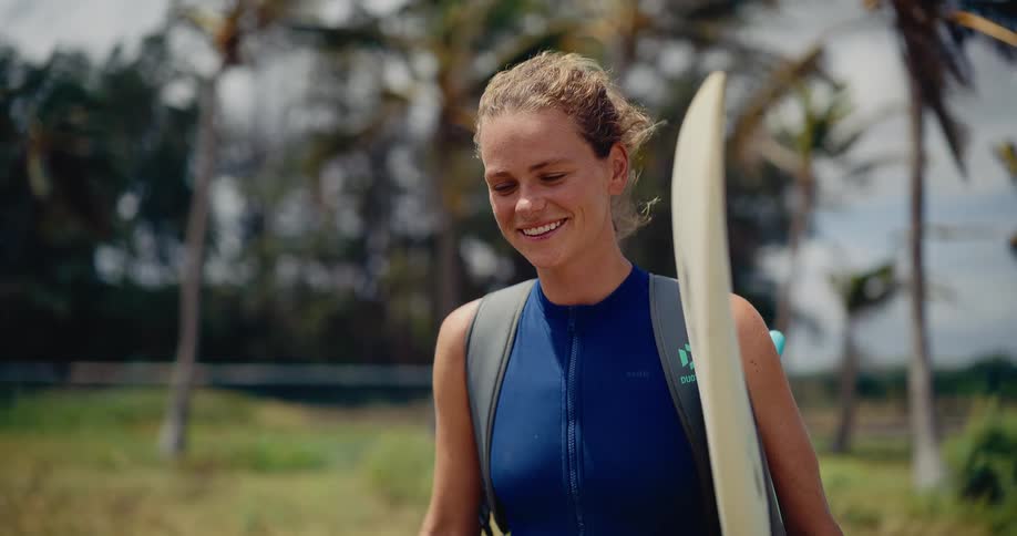 A happy young woman in a wetsuit smiles as she walks with her surfboard through a tropical landscape on a sunny day
