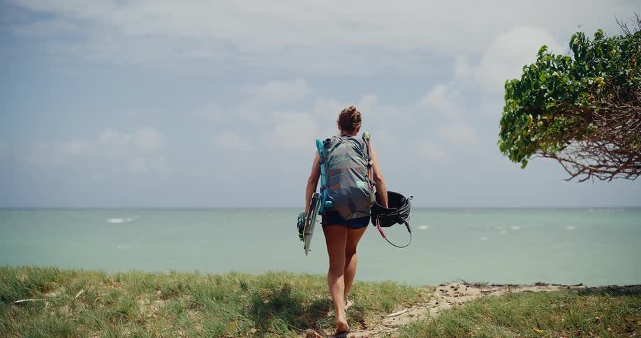 A female kitesurfer walks down a sandy path with her gear and stops to admire the beautiful ocean view