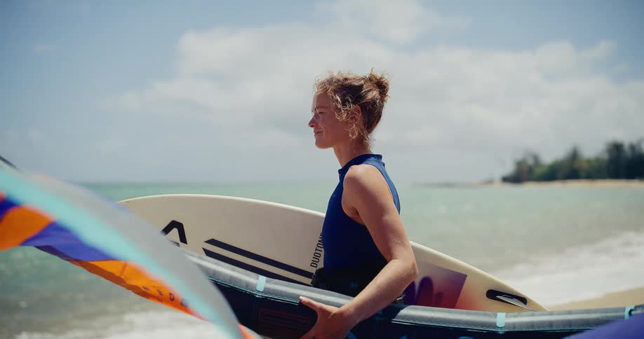 A smiling woman in slow motion holds her surfboard and wingfoil gear on a sunny beach, looking out at the ocean as the wing billows in the wind