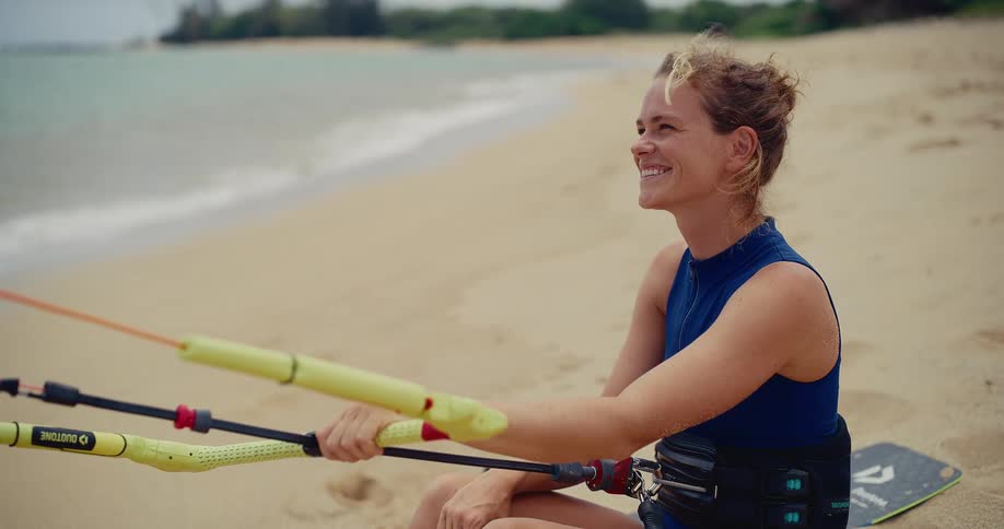 A female kitesurfer sits on a sandy beach, smiling happily as she holds her control bar and looks out at the ocean waves